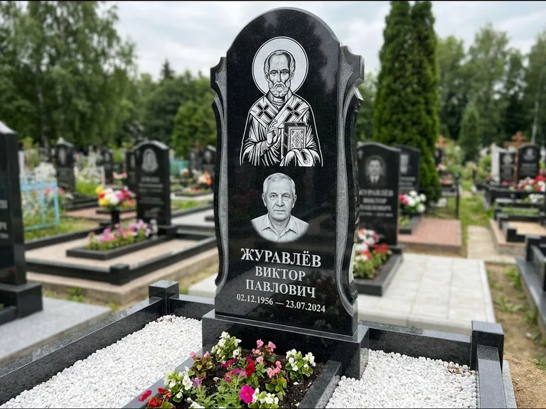 Памятник с гравировкой иконы: стела с образом в полный рост на обороте — гравировка образа Матроны Московской, низкие кусты спиреи в цвету, работа «Лебедева» в Краснодаре